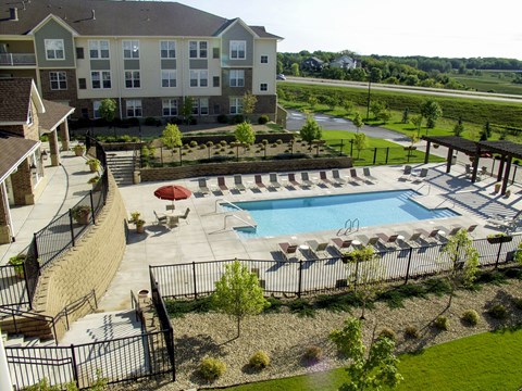 A sunlit outdoor pool here at Stoneleigh at the Reserve featuring a large rectangular swimming pool surrounded by a spacious concrete deck, rows of lounge chairs with small tables, and a shaded seating area with umbrellas, all enclosed by black metal fencing and framed by landscaped greenery, walking paths, and neighboring apartment buildings under open blue skies.