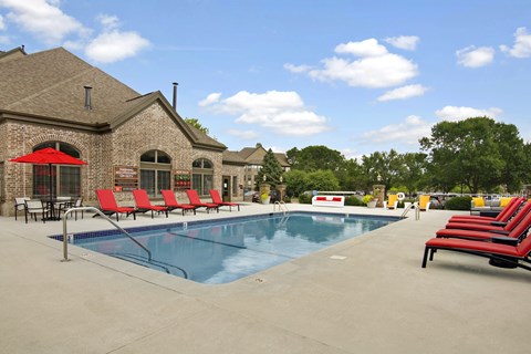 A resort-style outdoor swimming pool here at The Fairway featuring clear blue water with metal handrails, a wide concrete pool deck, and rows of red cushioned lounge chairs arranged for relaxation, alongside a brick clubhouse building with arched windows, shaded seating areas, mature trees, and open skies creating a welcoming community gathering space.