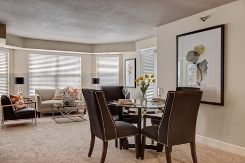 A welcoming living and dining area here at The Fairway showcasing a glass-top dining table with upholstered chairs, plush carpet flooring, and a comfortable seating area with a sofa, accent chair, and metallic coffee table, complemented by framed artwork, table lamps, and large windows with blinds that bring in soft natural light.