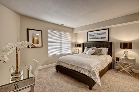 A serene bedroom here at The Fairway featuring a spacious layout with soft neutral carpeting, a plush bed with patterned bedding and layered pillows, matching bedside tables with decorative lamps, framed wall art, a large window with blinds that allows natural light, and a sleek desk area that adds both comfort and functionality.
