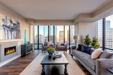 A stylish living room here at The Nic on Fifth showcasing rich wood-style flooring, a contemporary sofa and matching armchairs with accent pillows, and a dark wood coffee table centered on a light area rug, complemented by a sleek fireplace, modern wall art, greenery accents, and expansive floor-to-ceiling windows framing stunning city skyline views.