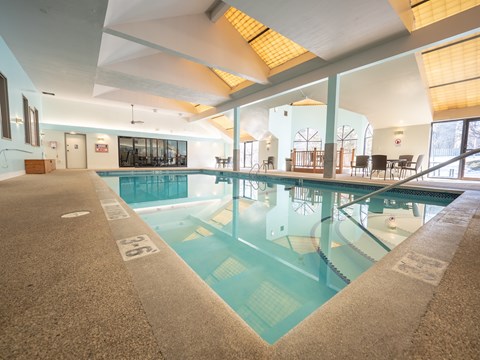 A sunny outdoor swimming pool here at Town Centre featuring a rectangular pool with clear blue water, stainless handrails, and a spacious concrete deck lined with lounge chairs, set against a backdrop of mature trees, open sky, and perimeter fencing that creates a relaxing space for warm-weather recreation.