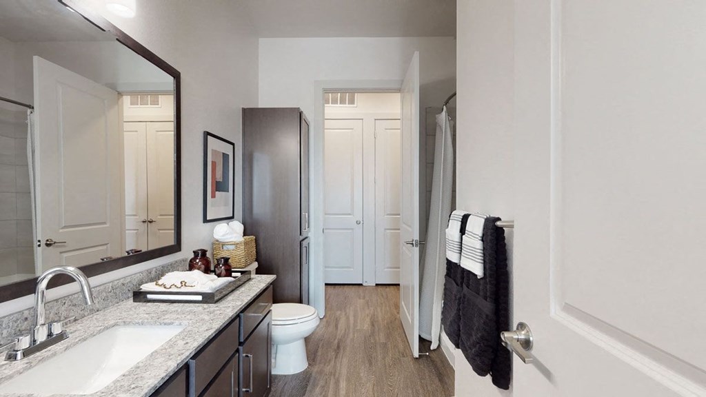 Modern bathroom here at Magnolia on the Green Apartment Homes with elegant gray granite countertop, large mirror, and dark wood cabinets. Warm wood floor, white walls, and striped towels create a sleek vibe.