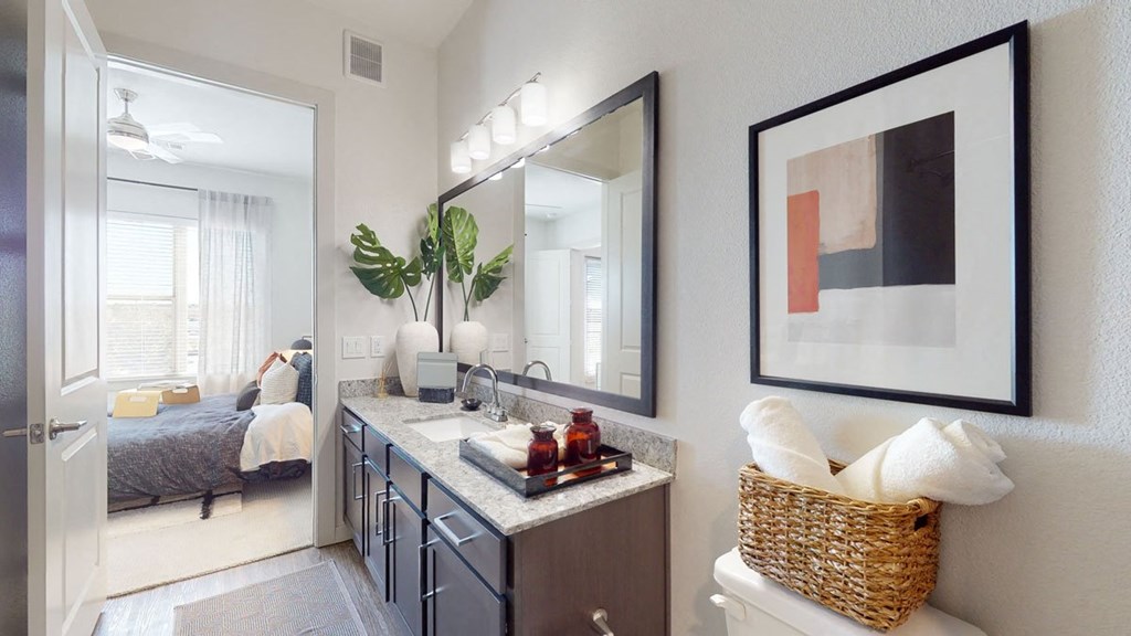 Modern bathroom here at Magnolia on the Green Apartment Homes with a granite countertop, dark cabinetry, and large mirror. Art on wall, basket with towels, and leafy plants add a fresh touch. Cozy bedroom visible through open door.