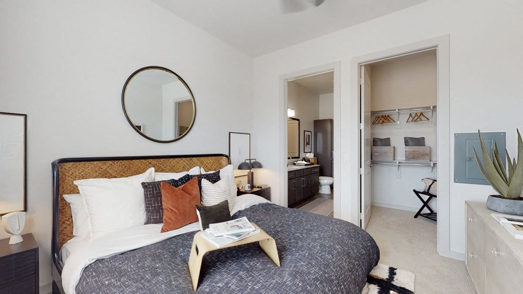 A cozy bedroom here at Magnolia on the Green Apartment Homes with a neatly made bed, colorful throw pillows, and a round mirror above the headboard. A bedside table has a lamp and decor. The open door reveals an ensuite bathroom with dark cabinets and a glimpse of a walk-in closet with hangers. The room conveys a modern and inviting atmosphere.