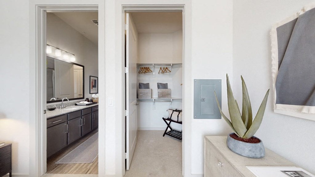 Modern bedroom interior here at Magnolia on the Green Apartment Homes with a view into a walk-in closet and bathroom. Neutral tones, a potted agave plant, and organized spaces create a calm, elegant atmosphere.