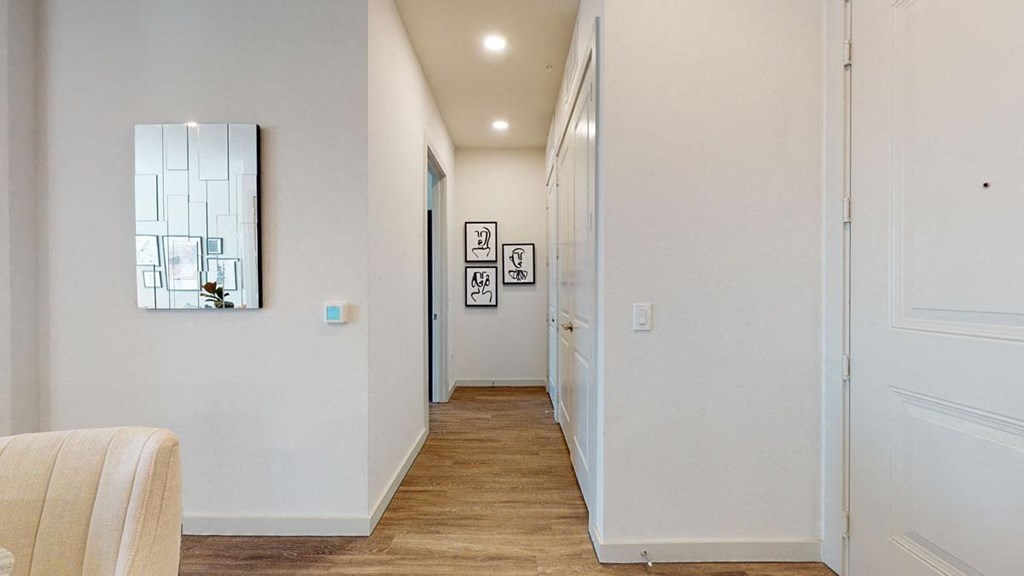 Modern hallway here at Magnolia on the Green Apartment Homes with wooden floor, neutral walls, and bright lighting. Features a mirrored wall panel, artwork, and a partially visible beige chair. Minimalist and inviting.