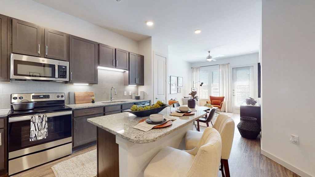 Modern kitchen and living area here at Magnolia on the Green Apartment Homes with wood cabinets, granite countertops, and a dining island. Cozy seating area with armchair, soft lighting, and large windows.