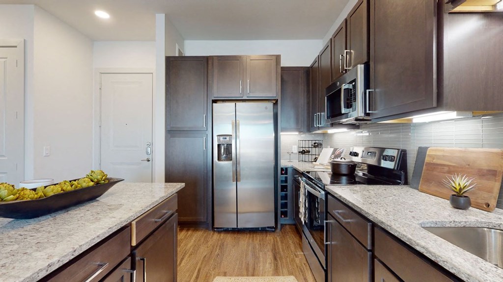 Modern kitchen here at Magnolia on the Green Apartment Homes with dark wood cabinets, stainless steel appliances, and granite countertops. Bowl of green apples adds a fresh touch. Bright, clean space.