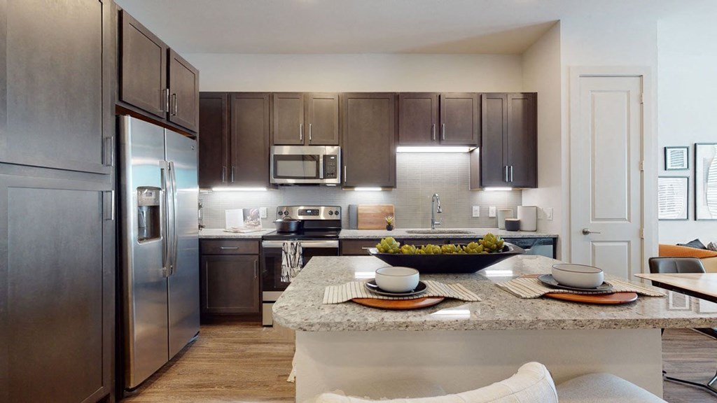 Modern kitchen here at Magnolia on the Green Apartment Homes with dark wood cabinets, stainless steel appliances, and a speckled granite island. Place settings and fruit bowl create a welcoming feel.
