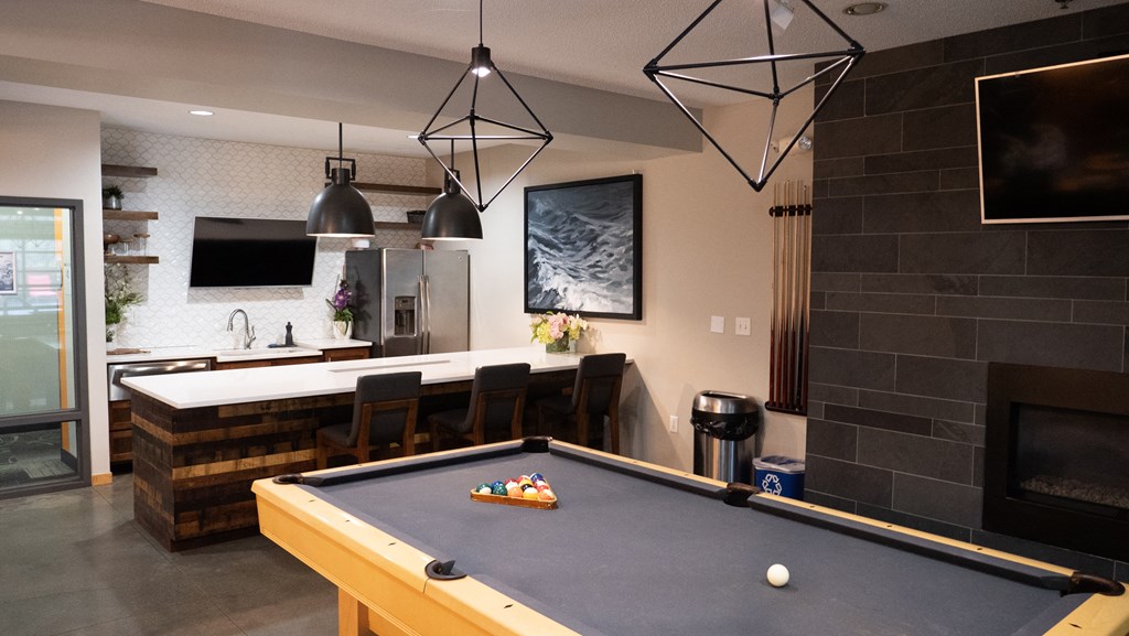 Modern game room here at Marbella on Dean Apartment Homes with a pool table, bar stools, and a kitchenette. Geometric pendant lights, abstract ocean art, and a cozy fireplace create a welcoming atmosphere.