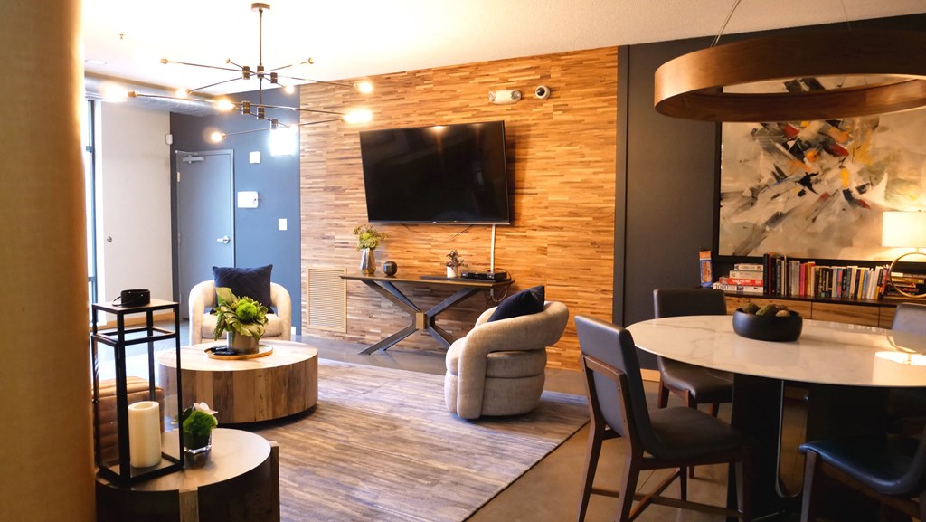 Modern living room here at Marbella on Dean Apartment Homes with wooden accents features a wall-mounted TV, circular table, abstract art, bookshelves, and plush seating, exuding warmth and style.