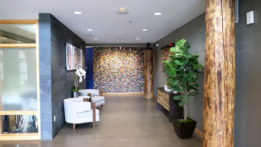 Modern office lobby here at Marbella on Dean Apartment Homes with gray armchairs, wooden columns, a green plant, and a colorful mosaic wall art. The atmosphere is warm and welcoming.