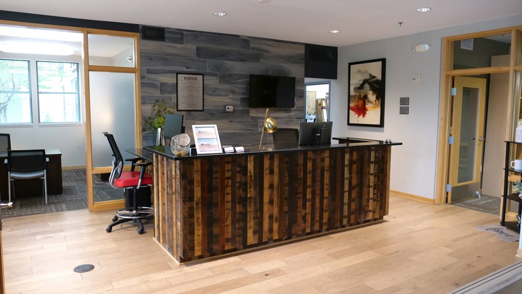 Modern office reception area here at Marbella on Dean Apartment Homes with a wooden desk, red and black chair, framed art, and glass partitions. Warm, welcoming atmosphere.