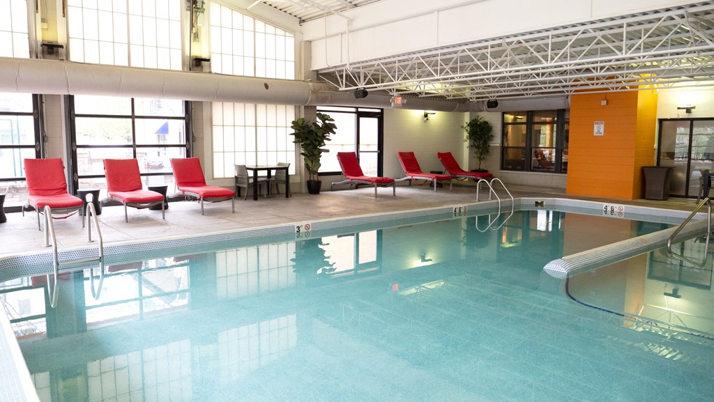 Indoor pool here at Marbella on Dean Apartment Homes with clear water, surrounded by red lounge chairs and potted plants. Large windows and an orange accent wall create a bright, inviting atmosphere.
