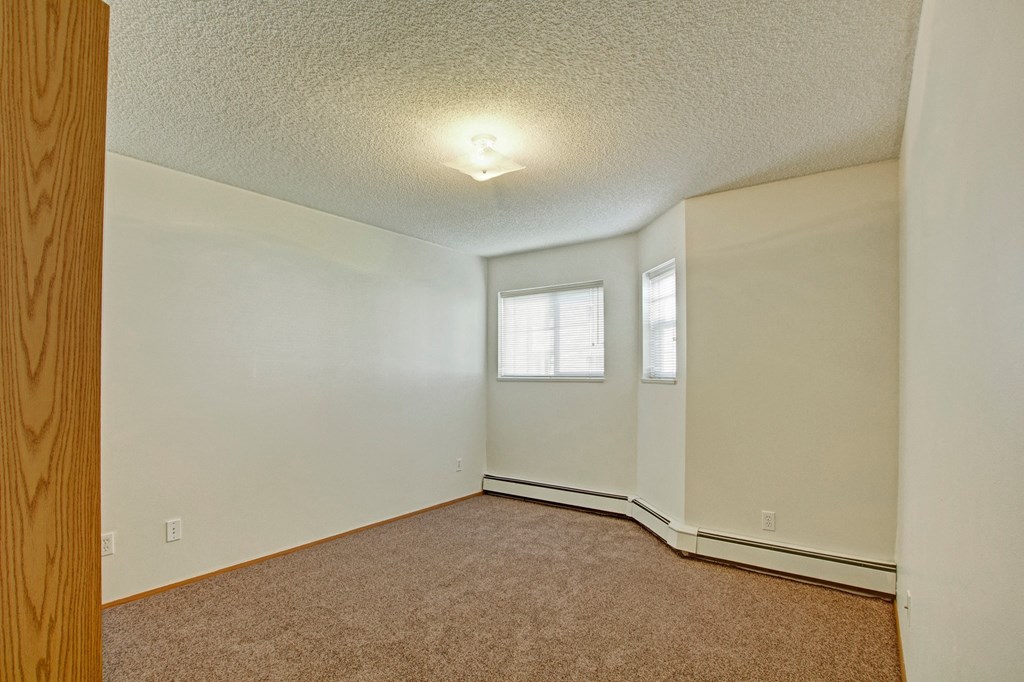 Empty bedroom here at Aspen Terrace Apartment Homes with beige carpet, cream walls, a small window with blinds, and a central ceiling light. The room feels clean and minimalistic.