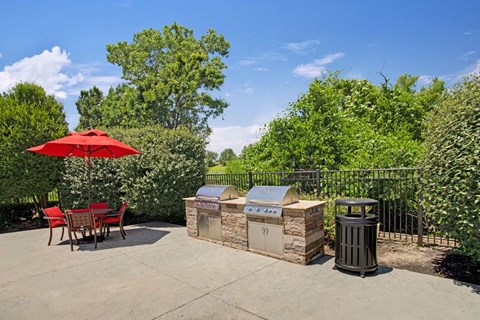 Outdoor patio here at Memorial Creek Apartment Homes with two stainless steel grills on a stone counter, a red umbrella over a round table with chairs, surrounded by lush green trees.