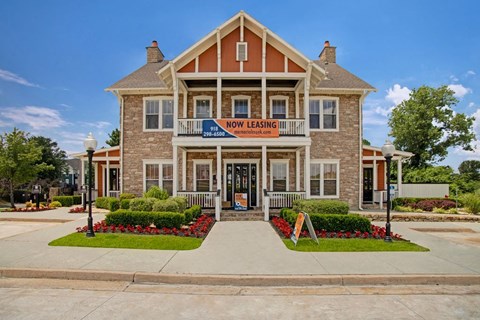 Two-story brick building here at Memorial Creek Apartment Homes with a "Now Leasing" banner on the balcony. Surrounded by manicured lawns, flowers, and street lamps under a clear blue sky.