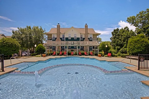 A serene outdoor pool here at Memorial Creek Apartment Homes with clear blue water, surrounded by red lounge chairs and greenery. A two-story house with chimneys is in the background, under a blue sky.