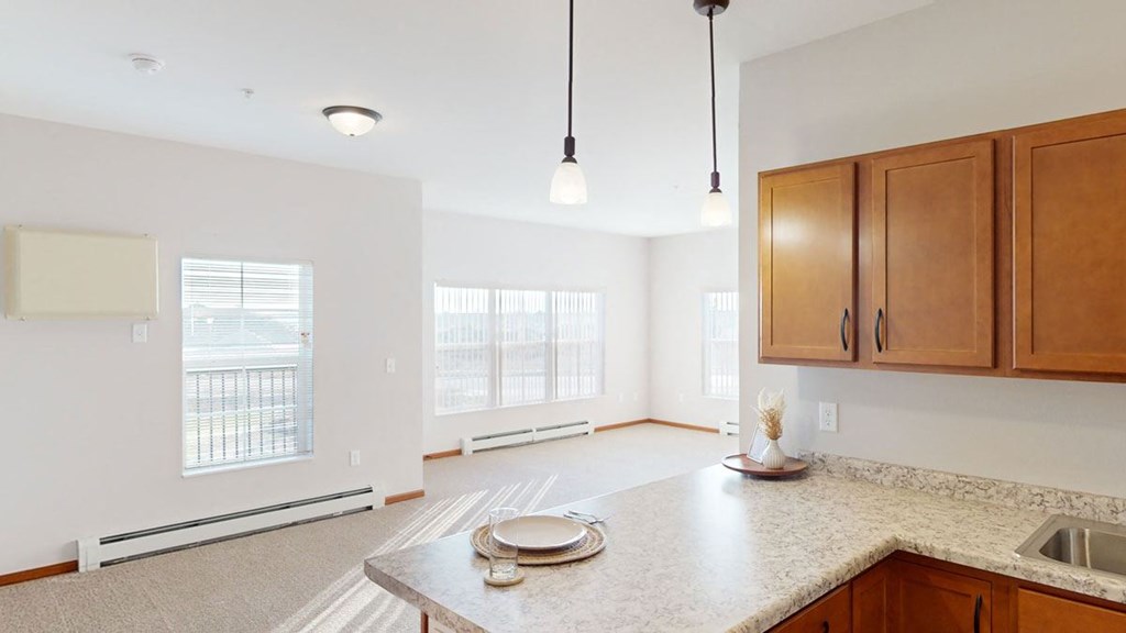 Bright open-plan kitchen and living area here at The Club at Heritage Apartment Homes with neutral walls. Features wooden cabinets, pendant lights, large windows, and light streaming in. Warm and inviting.