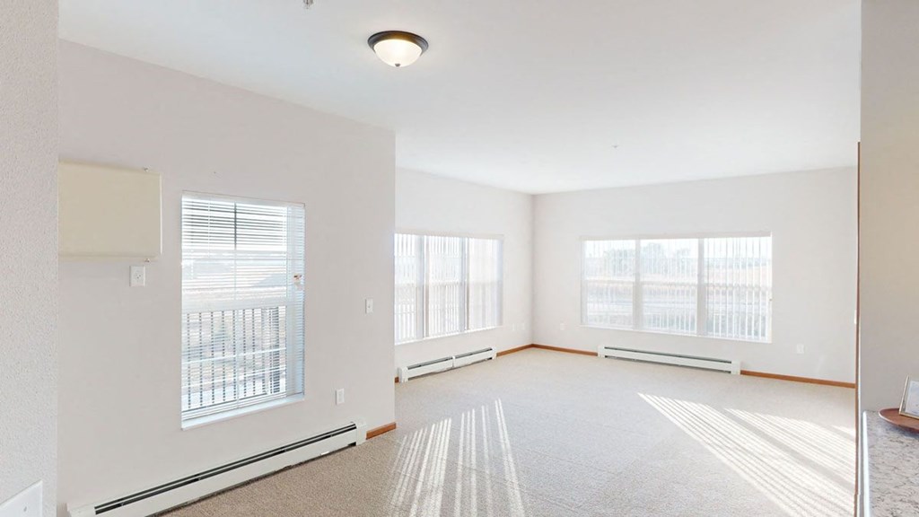 Spacious, sunlit room here at The Club at Heritage Apartment Homes with large windows and vertical blinds. Neutral-toned walls and carpet create a bright, airy atmosphere. Ceiling light adds warmth.