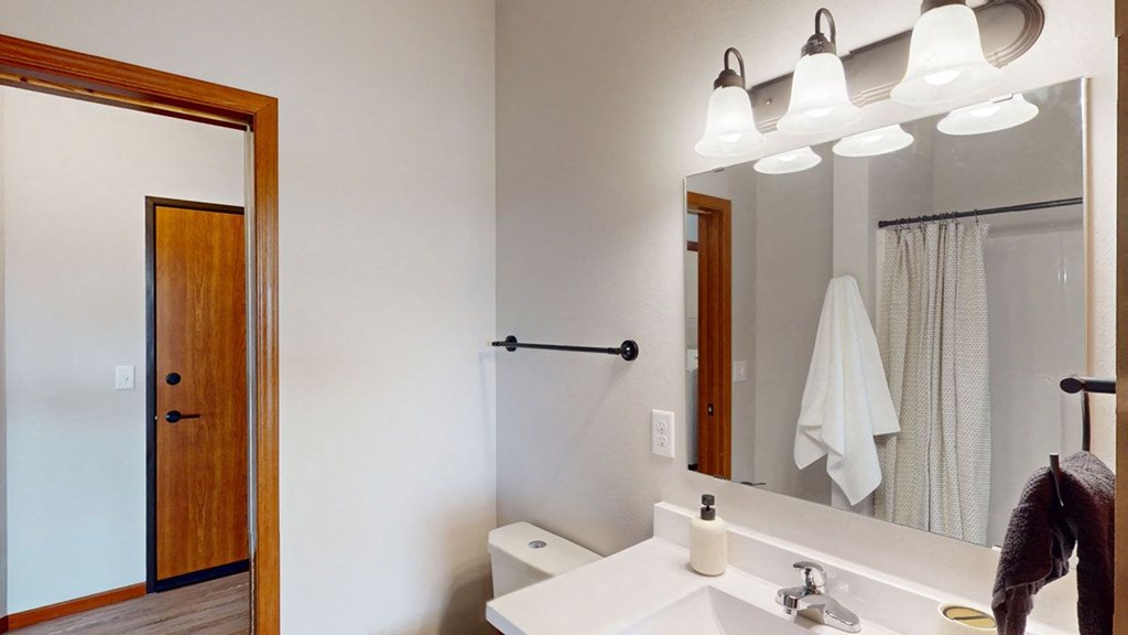 Bathroom here at The Club at Heritage Apartment Homes with a large mirror, three overhead lights, and wooden door. A towel hangs by the shower curtain. The sink holds a soap dispenser.