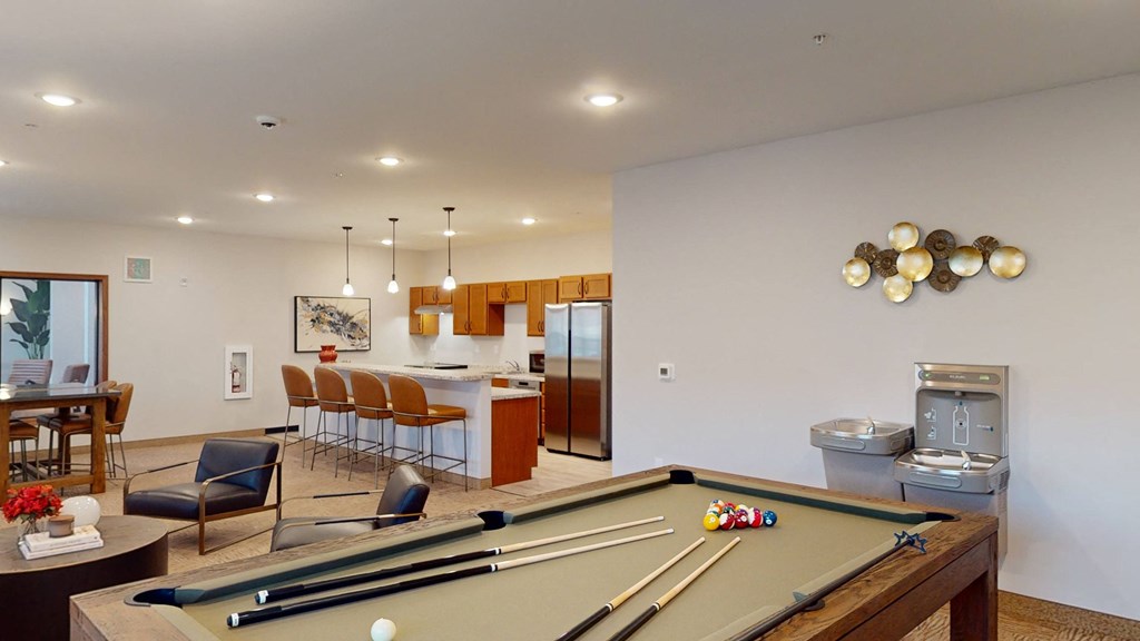 Stylish recreation room here at The Club at Heritage Apartment Homes with a pool table in the foreground, modern kitchen with barstools, chic decor, and a relaxed, inviting atmosphere.
