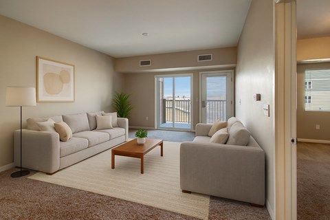 A bright living room here at Dakota Commons featuring two light neutral sofas arranged across a wooden coffee table, a soft area rug over carpeted flooring, neutral wall art, and a floor lamp, with sliding glass doors leading to a private balcony that brings in natural light and offers an open, comfortable layout.
