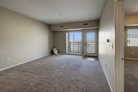 A spacious, unfurnished living room here at Dakota Commons featuring soft neutral walls, plush carpet flooring, and a sliding glass door with an adjacent exterior door that opens to a private balcony, allowing natural light to fill the open layout and highlight the room’s generous size.