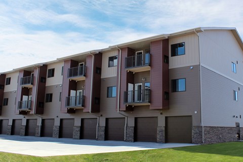 A modern apartment building exterior here at Dakota Commons featuring a three-story structure with private balconies, ground-level garages, mixed neutral siding with brick accents, and a well-maintained lawn in the foreground beneath a bright blue sky.