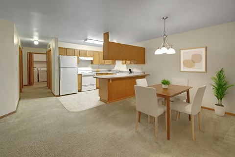 A spacious open-concept dining and kitchen area here at Dakota featuring warm wood cabinetry, a peninsula breakfast bar, white appliances, soft carpet transitioning to vinyl flooring, a four-seat dining table with modern chairs, and neutral lighting that creates a bright, welcoming apartment interior.