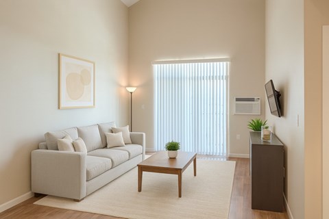 A bright, modern living room here at Dakota Ridge featuring high ceilings, a light neutral sofa with coordinating pillows, a wooden coffee table on a soft area rug, warm wood-style flooring, vertical blinds allowing natural light, a wall-mounted TV, and minimal decor creating an open, inviting atmosphere.