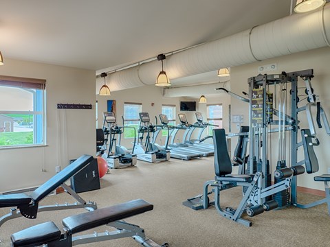 A spacious, well-lit fitness center here at Dakota Ridge featuring treadmills lined along large windows, a multifunction weight machine, a padded workout bench, neutral carpet flooring, exposed ceiling ductwork, and warm pendant lighting that creates a clean, motivating environment for residents to exercise comfortably.