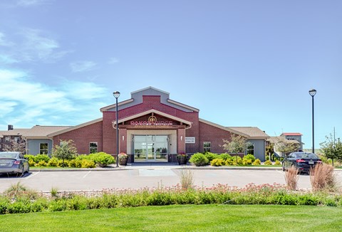 A welcoming exterior view here at Dakota Ridge featuring the brick clubhouse entrance with a peaked roofline, glass double doors, landscaped greenery, colorful planters, and a wide parking area set against a bright blue sky and well-maintained lawns.
