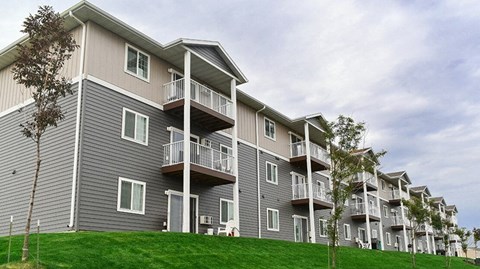 A modern apartment building exterior here at Pheasant Ridge showcasing multi-story residences with gray and beige siding, private balconies, white railings, evenly spaced windows, and a well-maintained green lawn with young trees under a lightly overcast sky.