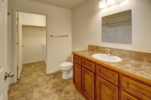 A clean neutral bathroom here at Phoenix Ridge featuring a wood vanity with a stone-look countertop and white sink, tile flooring in warm earth tones, a large wall mirror with overhead lighting, a toilet, and an adjacent walk-in closet with shelving and in-unit laundry access visible through the open doorway.