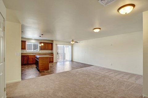 A spacious open living area here at Phoenix Ridge featuring soft neutral carpeting, an open-concept layout flowing into a kitchen with warm wood cabinetry, a center island, tile flooring, pendant lighting, and a sliding glass door that brings in natural light and connects to an outdoor space.