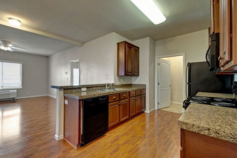 A bright open kitchen here at Prairie Vista featuring warm wood cabinetry, granite-style countertops, a double sink with breakfast bar seating, black appliances including a dishwasher and range, wood-look flooring, overhead lighting, and an open view into the living area with large windows bringing in natural light.
