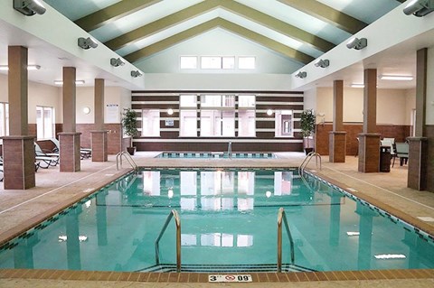 A bright indoor swimming pool here at Renaissance Heights featuring clear turquoise water, stainless steel handrails, a vaulted ceiling with exposed beams, large windows letting in natural light, tiled pool decking, and surrounding seating areas that create a relaxing, resort-style atmosphere.