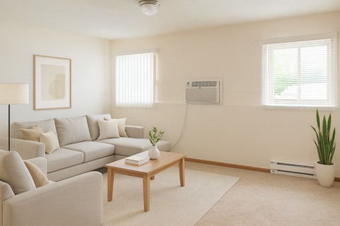 A bright neutral living room here at Rockridge featuring a light gray sectional sofa with matching throw pillows, a wooden coffee table on a soft area rug, beige walls, large windows with blinds providing natural light, and simple modern decor creating a clean and inviting apartment living space.