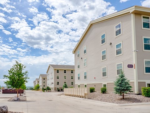 A well-maintained apartment community exterior here at Rockridge Apartments featuring beige multi-story buildings with white trim and multiple windows. A paved drive runs through the property, lined with mailboxes, landscaped rock beds, and small trees, creating an open, orderly setting under a bright, partly cloudy sky.