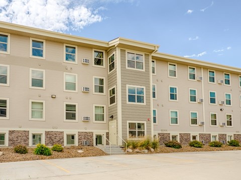 A modern, multi-story apartment building exterior here at Rockridge Apartments with beige siding, white trim, and rows of large windows. The building features a central entry with steps, landscaped rock beds with greenery along the base, and a paved parking area in front under a clear blue sky.