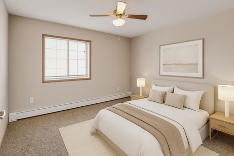 A calm, comfortable bedroom here at Roosevelt East Apartments with beige carpet, soft neutral walls, and a large window with blinds that lets in natural light. A ceiling fan with light hangs overhead, and the room is staged with a neatly made bed, matching nightstands, and lamps, creating a warm and relaxing atmosphere.