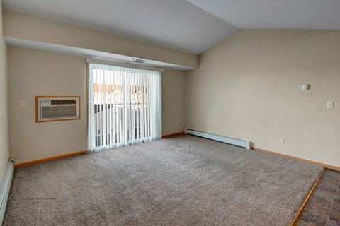 A spacious, empty living room here at Roosevelt East Apartments with beige carpet, soft neutral walls, and wood-trim baseboards. A sliding glass door with vertical blinds brings in natural light and opens to a patio area, while a wall-mounted air conditioning unit and vaulted ceiling add comfort and an open, airy feel.