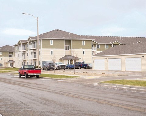 A well-kept apartment community exterior here at Roosevelt East Apartments featuring multi-story buildings with beige and green siding, white trim, and multiple balconies. A paved street runs alongside the buildings with parked vehicles, garage structures, and maintained lawns, creating a clean, residential neighborhood setting.