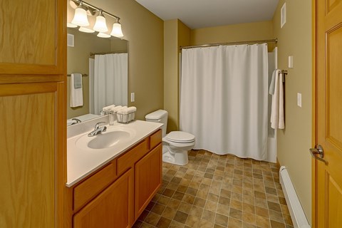 A clean, well-lit bathroom here at Vue 28 Apartments with tiled flooring, neutral walls, and a bathtub with a shower curtain. A wooden vanity with a white countertop and sink sits beneath a large wall mirror with overhead lighting, while a white toilet and towel bars complete the simple, functional layout.