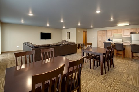 A spacious community gathering area here at Vue 28 Apartments featuring multiple wooden dining tables with chairs, a comfortable seating area with sofas, and an open kitchen with a breakfast bar and cabinetry. Recessed ceiling lights and patterned flooring create a bright, welcoming space designed for socializing, dining, or group activities.