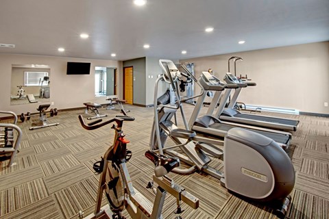 A well-equipped fitness center here at Vue 28 Apartments featuring cardio machines, weight equipment, and exercise benches arranged across a spacious, open floor plan. The room includes mirrored walls, wall-mounted televisions, patterned flooring, and bright overhead lighting, creating a comfortable and functional space for daily workouts.