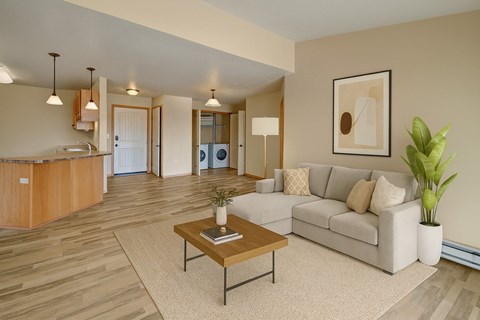 A bright, open living room here at Williston Garden Apartments with wood-style flooring, soft neutral walls, and an inviting layout that flows into the kitchen area. A light-colored sectional sofa sits on an area rug with a wooden coffee table, complemented by pendant lighting, simple wall art, and an in-home laundry area visible in the background.