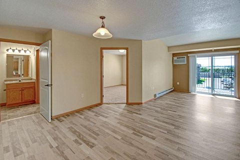 A spacious, open living area here at Williston Garden Apartments with wood-style flooring, soft neutral walls, and an airy layout. A sliding glass door with vertical blinds leads to a private balcony and allows natural light to fill the room, while baseboard heating, overhead lighting, and nearby access to the bedroom and bathroom add comfort and convenience.